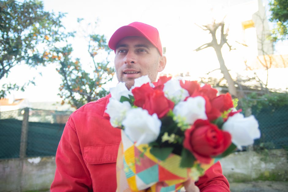 A delivery person dressed in a red cap and matching red jacket holds a vibrant bouquet of fresh flowers outdoors. The bouquet features red and white roses, with green foliage, wrapped in colorful pape