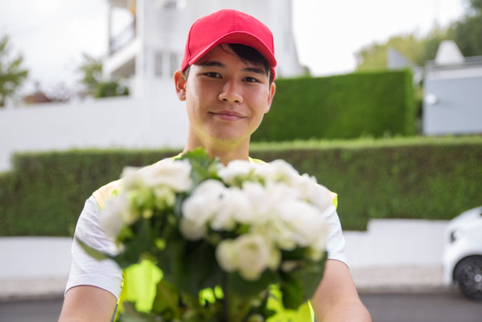 A smiling young delivery person with a red cap and high-visibility vest is holding a bouquet of fresh white roses and green foliage, offering the floral gift towards the camera. The bouquet is wrapped