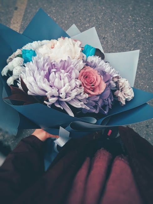 A vibrant floral bouquet featuring a large, white and lilac chrysanthemum centerpiece surrounded by pink roses, purple statice, and small blue flowers, all arranged in a tightly packed style. The bouq