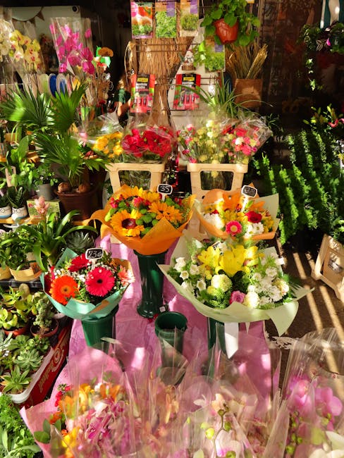 A vibrant display of fresh flower bouquets arranged on pink cloth in a garden centre or florist shop setting, showcasing a variety of blooms including bright orange gerbera daisies, pink chrysanthemum